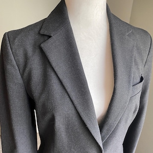 Op de afbeelding: Een antracietgrijze blazer met een revers en een sluiting met &eacute;&eacute;n knoop. De blazer heeft een getailleerde pasvorm, een borstzak en een klassiek ontwerp. De stof lijkt een geweven materiaal te zijn, geschikt voor professionele of formele kleding.