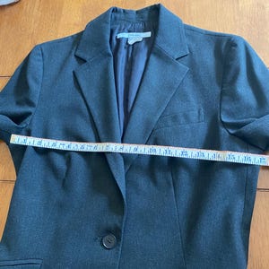 Op de afbeelding: Een donkergrijze blazer met een enkele knoopsluiting, een gekerfde revers en twee voorzakken. De blazer wordt gemeten met een meetlint. Het label luidt "ZARA BASIC".
