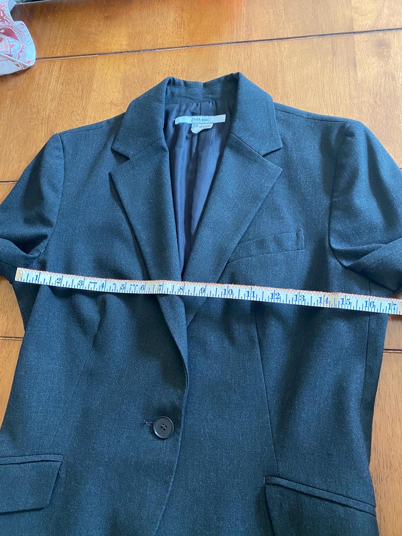 Op de afbeelding: Een donkergrijze blazer met een enkele knoopsluiting, een gekerfde revers en twee voorzakken. De blazer wordt gemeten met een meetlint. Het label luidt "ZARA BASIC".