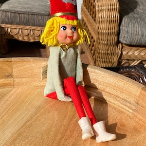 Puede incluir: Una muñeca de elfo de Navidad vintage con un gorro rojo, cabello amarillo y botas blancas. El elfo está sentado en una superficie de madera.
