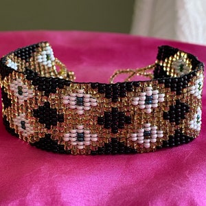 May include: A black and gold beaded bracelet with white flower-shaped beads. The bracelet is made with small, tightly woven beads.