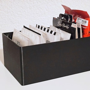 Könnte beinhalten: Eine schwarze Holzkiste gefüllt mit Gitarrensaiten. Die Kiste enthält verschiedene Saitenpackungen, darunter Dean Markley und andere Marken. Die Saiten sind in weißer und schwarzer Verpackung, mit etwas roter Verpackung sichtbar.