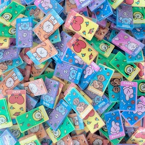 May include: A collection of colorful rectangular charms featuring cartoon characters in various poses. The charms are in shades of blue, green, yellow, orange, and purple. Some charms have text like "COOKY" and "TATA".