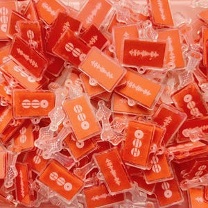 May include: A collection of small, rectangular orange acrylic charms featuring white printed designs. Some charms display a horizontal sound wave graphic, while others show a circular icon with three horizontal lines. Each piece has a clear loop.