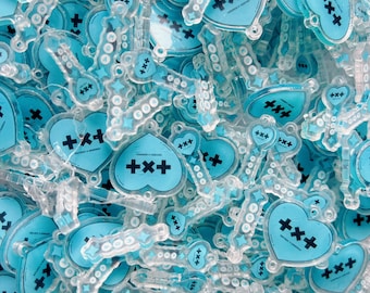 May include: A collection of clear acrylic charms featuring a light blue heart shape with a black plus sign logo and the text TOMORROW X TOGETHER. Each charm includes a small loop for attachment and decorative star accents on the surface.