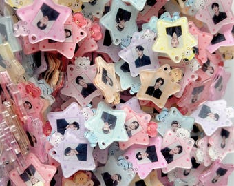 May include: A collection of star-shaped acrylic charms in pastel colors, each featuring a small portrait of a person in the center. The charms are translucent and layered, with small character figures attached to the edges of the star frames.