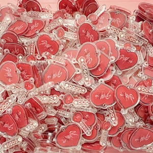 May include: A collection of clear acrylic keychains with a red heart design and the word "Stray" in white script. The keychains are in various shapes, including hearts and rectangular bars, and are scattered on a pink surface.