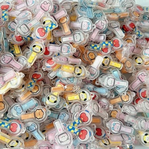 May include: A large collection of small, clear acrylic charms shaped like light sticks, each featuring a colorful cartoon character face. The charms include various pastel and bright colors like yellow, blue, pink, and red, with small loops for attachment.