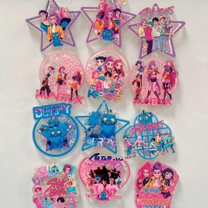 May include: A collection of colorful, glitter-filled acrylic charms featuring K-pop group illustrations and the text "Derpy," "Huntrix," and "Girl Boys." The charms are in various shapes, including stars, circles, and hearts.