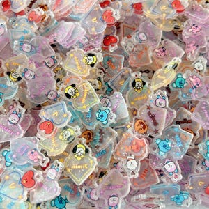 May include: A collection of small, translucent acrylic charms featuring colorful cartoon characters and star motifs. Each charm has a clear, glossy finish with printed designs in shades of blue, pink, yellow, and red, scattered in a dense pile.