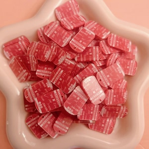 May include: A collection of small, red rectangular tags with rounded corners, each with white text and a hole for attachment. The tags are piled in a light pink, star-shaped dish, set against a matching pink background.