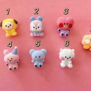 May include: A collection of seven colorful, cartoon-style figurines on a pink background. The figurines include animal and character designs in various colors like yellow, pink, blue, and white. Each figurine has unique features and expressions.