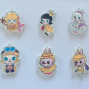 May include: A collection of six cartoon character charms with a clear, glossy finish. The charms feature various figures, including a baby, a girl with a crown, and a bunny-like character, each with unique outfits and accessories. The color palette includes yellow, blue, and pink.