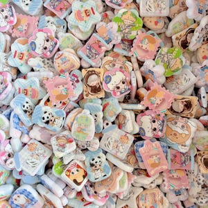 May include: A collection of colorful, cartoon-style charms in various shapes, including stars, rectangles, and flowers. The charms feature cute animal and character designs in pastel colors like pink, blue, and green. Some have text.
