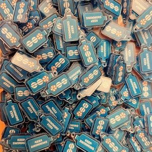 May include: A collection of small, blue, bottle-shaped acrylic charms. Each charm features a white graphic of a life preserver and the text "KEEP SWIMMING" printed in white. The charms are flat, rectangular with rounded edges, and have a small hole at the top for attachment.
