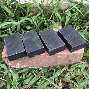 Può includere: Quattro saponette rettangolari grigio scuro sono disposte fianco a fianco su un mattone rosso. Le saponette sono su un letto di erba verde. Le saponette sembrano fatte a mano.