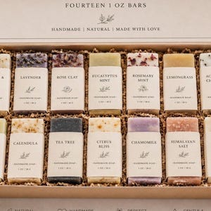 May include: A cardboard gift box containing fourteen 1 oz handmade soap bars, each wrapped in a paper label. The labels are printed with the names Oatmeal & Honey, Lavender, Rose Clay, Eucalyptus Mint, Rosemary Mint, Lemongrass, Activated Charcoal, Green Tea, Calendula, Tea Tree, Citrus Bliss, Chamomile, Himalayan Salt, and Coconut Milk.