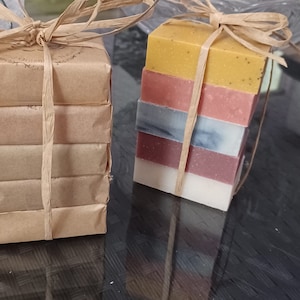 May include: Two stacks of handmade soap bars wrapped in brown paper and tied with twine. The top stack has five bars in shades of yellow, pink, blue, and white. The bottom stack has four bars wrapped in brown paper.