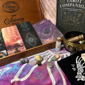 May include: An assortment of spiritual items including tarot cards, a book titled "Tarot Companion", a singing bowl, crystals, and a wooden box. The cards feature celestial and atmospheric imagery. A small bottle contains white stones.