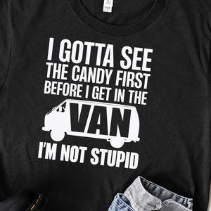 May include: Black t-shirt with white text that reads "I GOTTA SEE THE CANDY FIRST BEFORE I GET IN THE VAN I'M NOT STUPID." The shirt also features a white graphic of a van. The shirt is paired with blue jeans and black shoes.