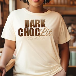 May include: Cream-colored t-shirt with the text "DARK CHOC Lit 100% Melanin" in brown lettering. The shirt has short sleeves and a relaxed fit. The person is wearing pink pants.