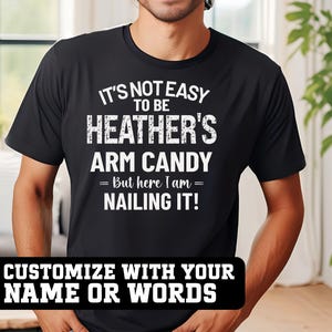 May include: Black t-shirt with white text that reads "IT'S NOT EASY TO BE HEATHER'S ARM CANDY = But here I am = NAILING IT!". The bottom of the shirt has a black banner that says "CUSTOMIZE WITH YOUR NAME OR WORDS".