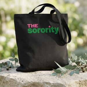 May include: Black tote bag with the text "THE Sorority" in pink and green. The bag has two black handles and is sitting on a stone surface with greenery in the background.