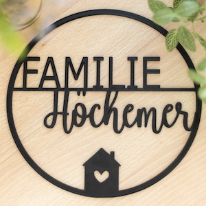 K&ouml;nnte beinhalten: Ein kreisf&ouml;rmiges schwarzes Wandschild mit dem Wort FAMILIE in Blockbuchstaben &uuml;ber dem Wort H&ouml;chemer in Schreibschrift. Unter dem Text befindet sich eine kleine Haussilhouette mit einem herzf&ouml;rmigen Ausschnitt in der Mitte.