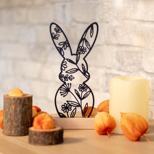 Könnte beinhalten: Schwarze Metall-Hasen-Silhouette mit floralen Ausschnitten auf einem Holzsockel. Der Hase ist von dekorativen Elementen umgeben, darunter eine weiße Kerze, kleine Holzblöcke und orangefarbene Trockenblumen, die eine saisonale Dekoration bilden.