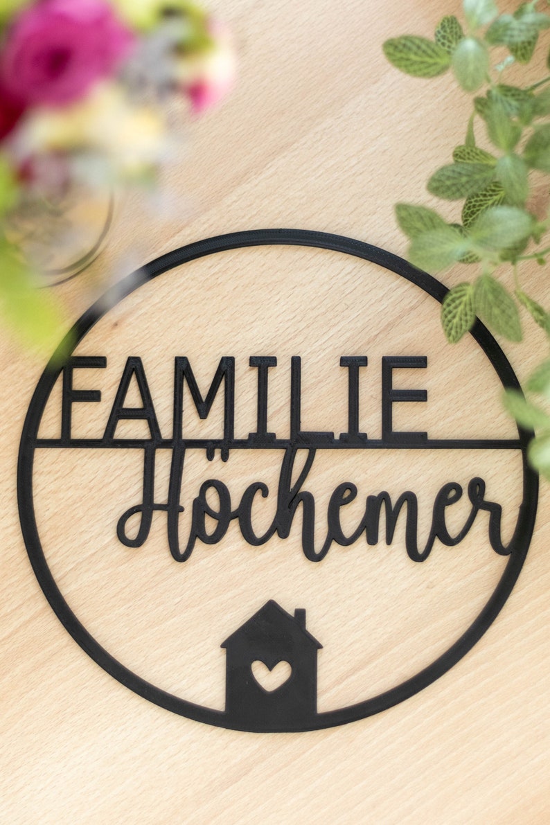 K&ouml;nnte beinhalten: Ein kreisf&ouml;rmiges schwarzes Wandschild mit dem Wort FAMILIE in Blockbuchstaben &uuml;ber dem Wort H&ouml;chemer in Schreibschrift. Unter dem Text befindet sich eine kleine Haussilhouette mit einem herzf&ouml;rmigen Ausschnitt in der Mitte.