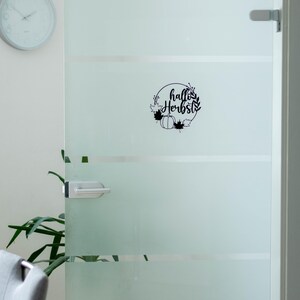 Op de afbeelding: Een zwarte vinylsticker aangebracht op een deur van matglas. Het ontwerp toont een cirkelvormige krans van bladeren en een pompoen, met de tekst "hallo Herbst" in cursief schrift in het midden van de cirkelvormige opstelling.