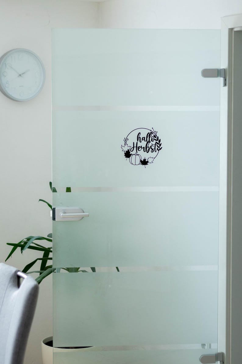 Op de afbeelding: Een zwarte vinylsticker aangebracht op een deur van matglas. Het ontwerp toont een cirkelvormige krans van bladeren en een pompoen, met de tekst "hallo Herbst" in cursief schrift in het midden van de cirkelvormige opstelling.