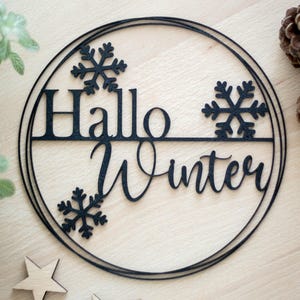 Könnte beinhalten: Eine kreisförmige schwarze Wanddekoration mit dem Schriftzug "Hallo Winter" in einer Kombination aus Serifenschrift und Schreibschrift. Das Design enthält drei sechsstrahlige Schneeflocken-Ausschnitte, die in den Rahmen aus zwei dünnen Ringen integriert sind.