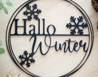 Op de afbeelding: Een ronde zwarte wanddecoratie met de tekst "Hallo Winter" in een combinatie van schreef- en scriptlettertypes. Het ontwerp is voorzien van drie zwarte uitgesneden sneeuwvlokken binnen de cirkelvormige lijst.
