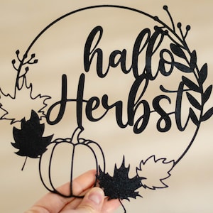 Op de afbeelding: Een zwart, cirkelvormig decoratief object met de tekst "hallo Herbst" in een sierlijk lettertype. Het ontwerp bevat de omtrek van een pompoen, diverse bladeren in de vorm van esdoornbladeren en dunne takjes langs de rand.