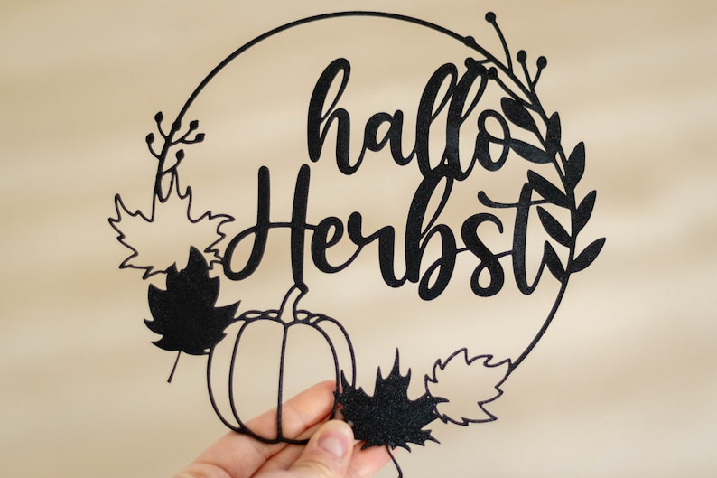 Op de afbeelding: Een zwart, cirkelvormig decoratief object met de tekst "hallo Herbst" in een sierlijk lettertype. Het ontwerp bevat de omtrek van een pompoen, diverse bladeren in de vorm van esdoornbladeren en dunne takjes langs de rand.