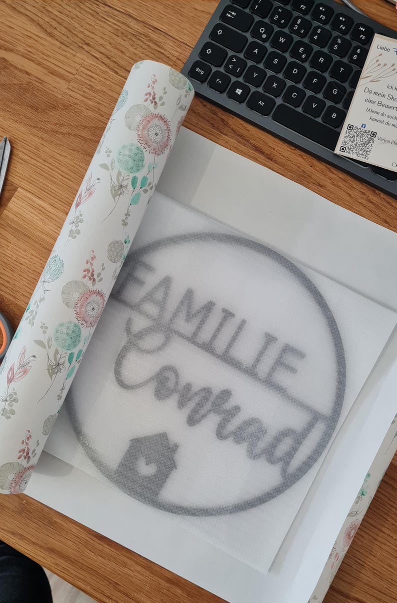 K&ouml;nnte beinhalten: Ein kreisf&ouml;rmiges graues Wandschild mit dem Text FAMILIE Conrad und einem kleinen Haussymbol, platziert auf wei&szlig;em Schutzpapier neben einer Rolle Geschenkpapier mit einem Muster aus rosa und gr&uuml;nen Bl&uuml;tenkugeln und Bl&auml;ttern.
