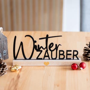 Könnte beinhalten: Eine winterliche Dekoration mit einem schwarzen Holzschild "Winter Zauber", einem kleinen Haus, Tannenzapfen und roten Kugeln. Alles steht auf einer hellen Holzoberfläche und schafft eine festliche Atmosphäre.