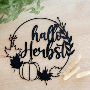 Op de afbeelding: Een zwarte cirkelvormige wanddecoratie met de tekst "hallo Herbst" in een cursief lettertype. Het ontwerp bevat een pompoen, diverse esdoornbladeren en dunne takjes die in de ronde omlijsting zijn verwerkt.