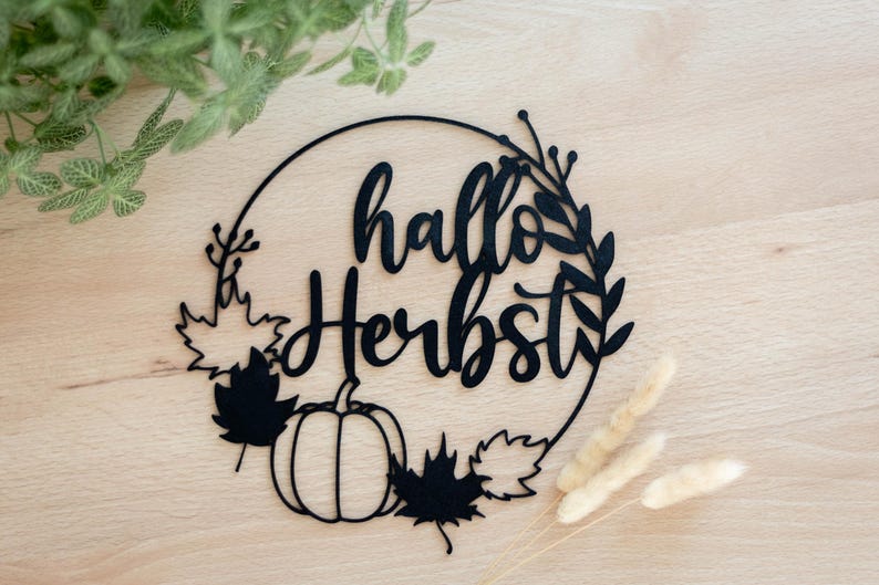 Op de afbeelding: Een zwarte cirkelvormige wanddecoratie met de tekst "hallo Herbst" in een cursief lettertype. Het ontwerp bevat een pompoen, diverse esdoornbladeren en dunne takjes die in de ronde omlijsting zijn verwerkt.