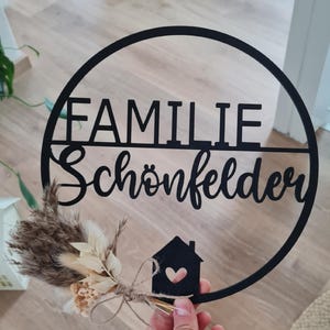 K&ouml;nnte beinhalten: Ein kreisf&ouml;rmiges schwarzes Wandschild mit dem Schriftzug "FAMILIE Sch&ouml;nfelder" in Block- und Schreibschrift. Ein kleines schwarzes Haus-Symbol mit Herz-Ausschnitt ist angebracht und mit getrockneten beigen und braunen Gr&auml;sern verziert.