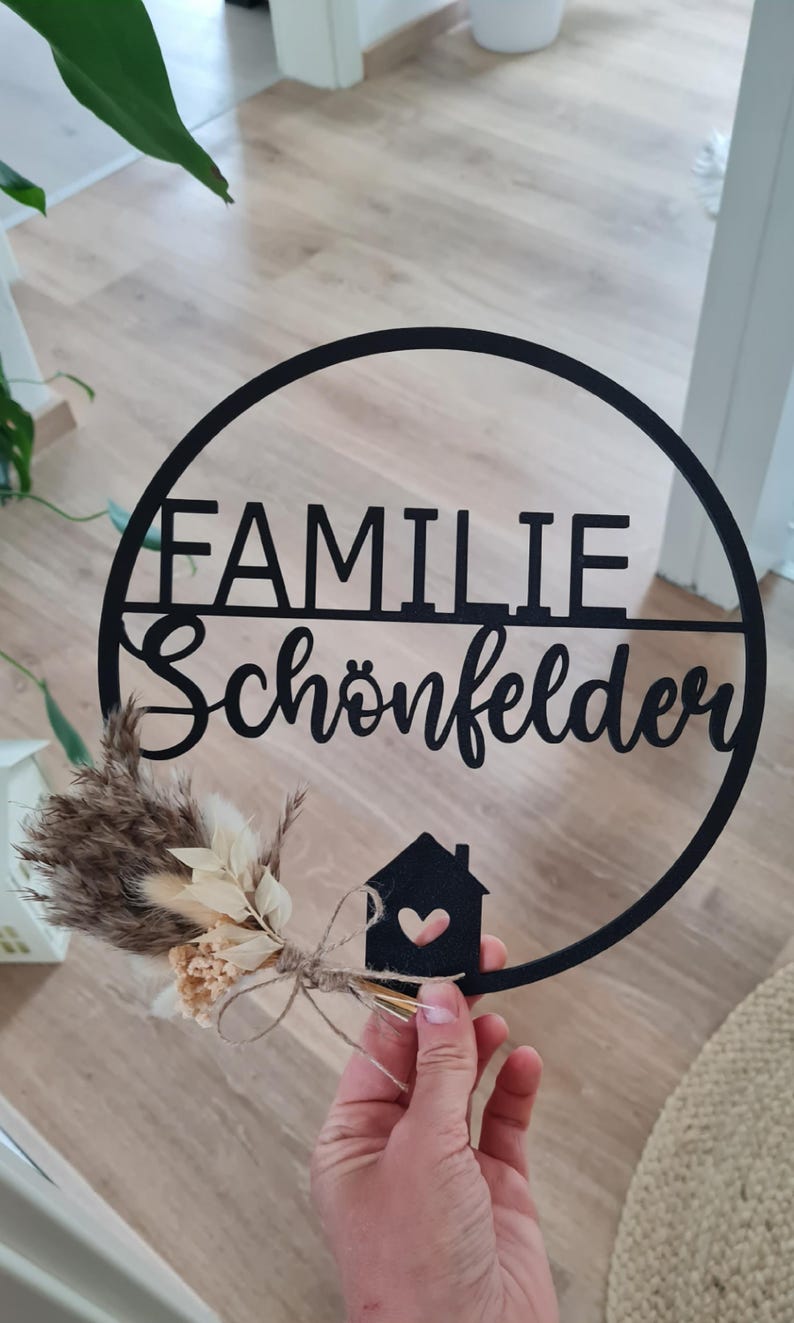 K&ouml;nnte beinhalten: Ein kreisf&ouml;rmiges schwarzes Wandschild mit dem Schriftzug "FAMILIE Sch&ouml;nfelder" in Block- und Schreibschrift. Ein kleines schwarzes Haus-Symbol mit Herz-Ausschnitt ist angebracht und mit getrockneten beigen und braunen Gr&auml;sern verziert.