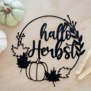 Op de afbeelding: Een zwarte cirkelvormige wanddecoratie met de tekst "hallo Herbst" in een cursief lettertype, omringd door silhouetten van esdoornbladeren, een pompoen en takken. Twee kleine decoratieve pompoenen en gedroogde grasstengels liggen ernaast.