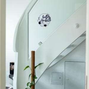 Op de afbeelding: Een zwarte, ronde muursticker met de tekst "hallo Herbst" in een sierlijk lettertype, omringd door een kransontwerp van bladeren, takken en een pompoen. De sticker is aangebracht op een wit glazen paneel van een trapleuning.