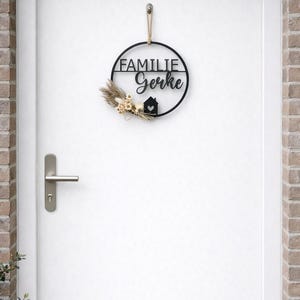 K&ouml;nnte beinhalten: Ein schwarzes, kreisf&ouml;rmiges Wandschild mit der Aufschrift FAMILIE Gerke, einem kleinen Haussymbol mit Herz sowie einem Arrangement aus getrockneten beigen Blumen und Gr&auml;sern. Das Schild h&auml;ngt an einer hellbraunen Kordel an einer wei&szlig;en T&uuml;r.