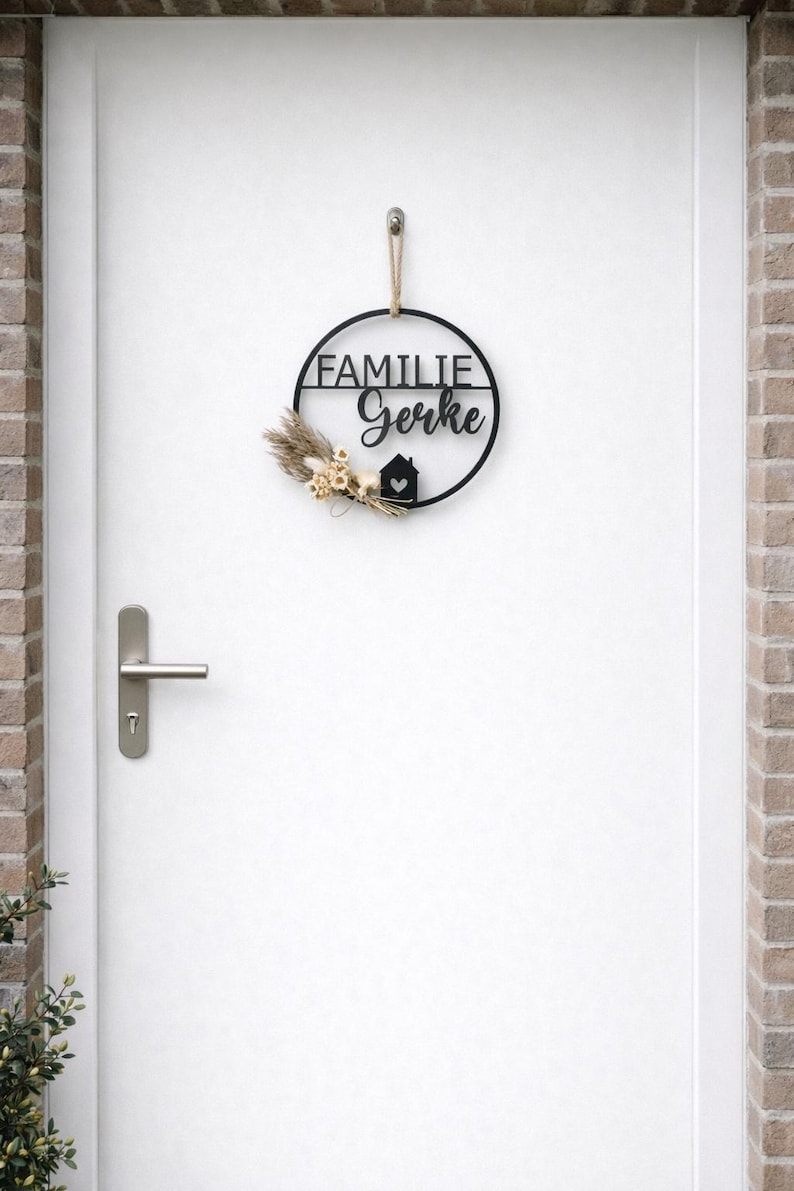K&ouml;nnte beinhalten: Ein schwarzes, kreisf&ouml;rmiges Wandschild mit der Aufschrift FAMILIE Gerke, einem kleinen Haussymbol mit Herz sowie einem Arrangement aus getrockneten beigen Blumen und Gr&auml;sern. Das Schild h&auml;ngt an einer hellbraunen Kordel an einer wei&szlig;en T&uuml;r.