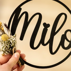 Könnte beinhalten: Ein schwarzes kreisförmiges Schild mit dem Namen Milo in Schreibschrift und einem kleinen Herz über dem Buchstaben i, begleitet von einem kleinen Strauß aus getrockneten weißen, gelben und braunen Blumen, der mit Bindfaden am Kreis befestigt ist.