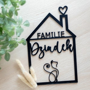Könnte beinhalten: Ein schwarzes Wandschild in Hausform mit dem Schriftzug "FAMILIE" und "Dziadek" in Schreibschrift. Das Design zeigt einen Schornstein mit einem Herz-Umriss und eine durchgehende Linienzeichnung einer sitzenden Katze unten.