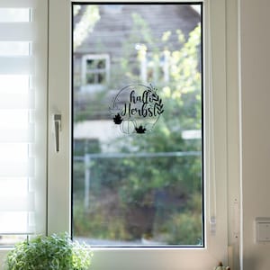 Op de afbeelding: Een zwarte ronde raamsticker met de tekst "hallo Herbst" in cursief schrift, omringd door een kransontwerp met een pompoen en esdoornbladeren. De sticker is aangebracht in het midden van een glazen raam.