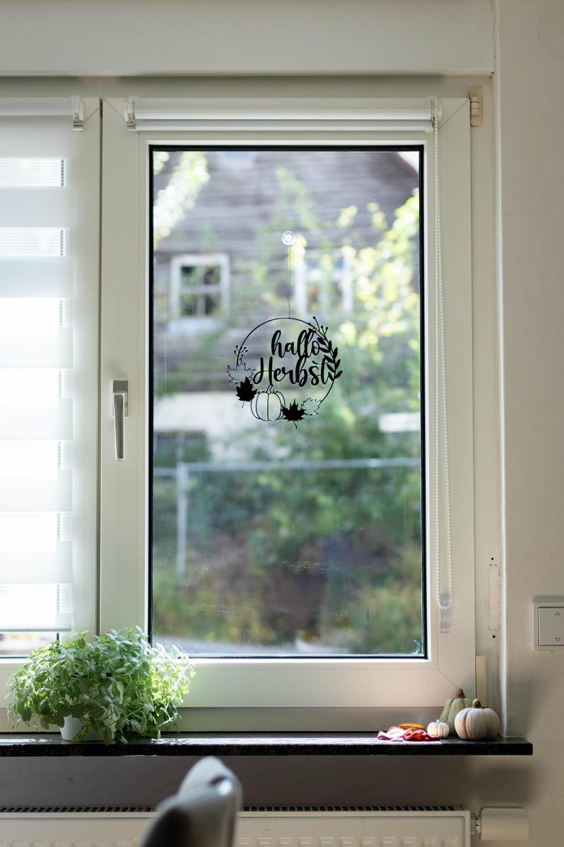 Op de afbeelding: Een zwarte ronde raamsticker met de tekst "hallo Herbst" in cursief schrift, omringd door een kransontwerp met een pompoen en esdoornbladeren. De sticker is aangebracht in het midden van een glazen raam.